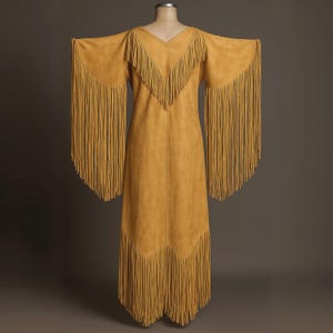 May include: Mustard-colored suede dress with long fringe detailing. The dress features a V-neckline and long sleeves with fringe. The hemline also has fringe. The dress is displayed on a mannequin against a neutral background.