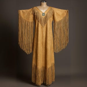 May include: A tan suede dress with long fringe sleeves and hem. The dress has a V-neckline with a turquoise and silver star-shaped embellishment. The dress is displayed on a mannequin against a neutral background.