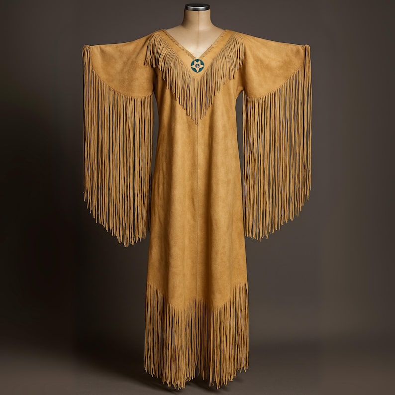 May include: A tan suede dress with long fringe sleeves and hem. The dress has a V-neckline with a turquoise and silver star-shaped embellishment. The dress is displayed on a mannequin against a neutral background.