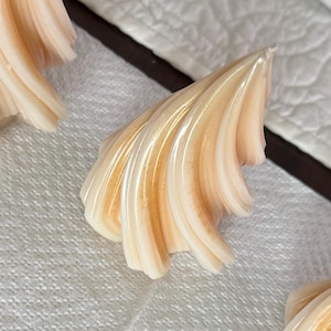 May include: Close-up of several seashell-shaped confections. The treats have a creamy white base with a golden-brown center and are arranged on a white, textured surface. The confections have a layered, wavy design.