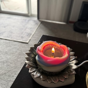 May include: A pink, blue, and white candle with a flame burning inside sits on a silver sunflower-shaped holder.