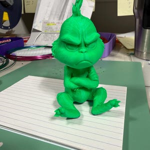 May include: A bright green 3D printed figurine of a character with a pointed head, furrowed eyebrows, and a grumpy facial expression. The figure is depicted in a seated position with arms crossed over its chest, featuring textured fur details.