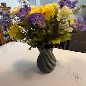 May include: A vibrant floral arrangement in a decorative vase. The bouquet features yellow, purple, and white flowers with green foliage. The vase has a unique, swirling design and is placed on a white surface.