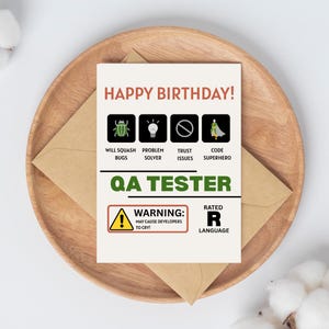 Può includere: Un biglietto di compleanno con la scritta "HAPPY BIRTHDAY!" e icone che rappresentano le competenze di un tester QA. La carta include un segnale di avvertimento e la scritta "QA TESTER". Poggia su un piatto di legno con una busta marrone.