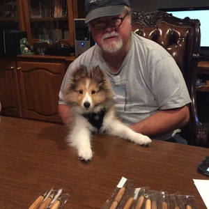 Puede incluir: Una colecci&oacute;n de bol&iacute;grafos de madera con detalles dorados, empaquetados en fundas de pl&aacute;stico transparente, se exhibe sobre una mesa de madera marr&oacute;n oscuro. Un perro peque&ntilde;o, esponjoso, marr&oacute;n y blanco est&aacute; sentado en la mesa. Un hombre con una camiseta gris y una gorra Just Golf est&aacute; en el fondo.