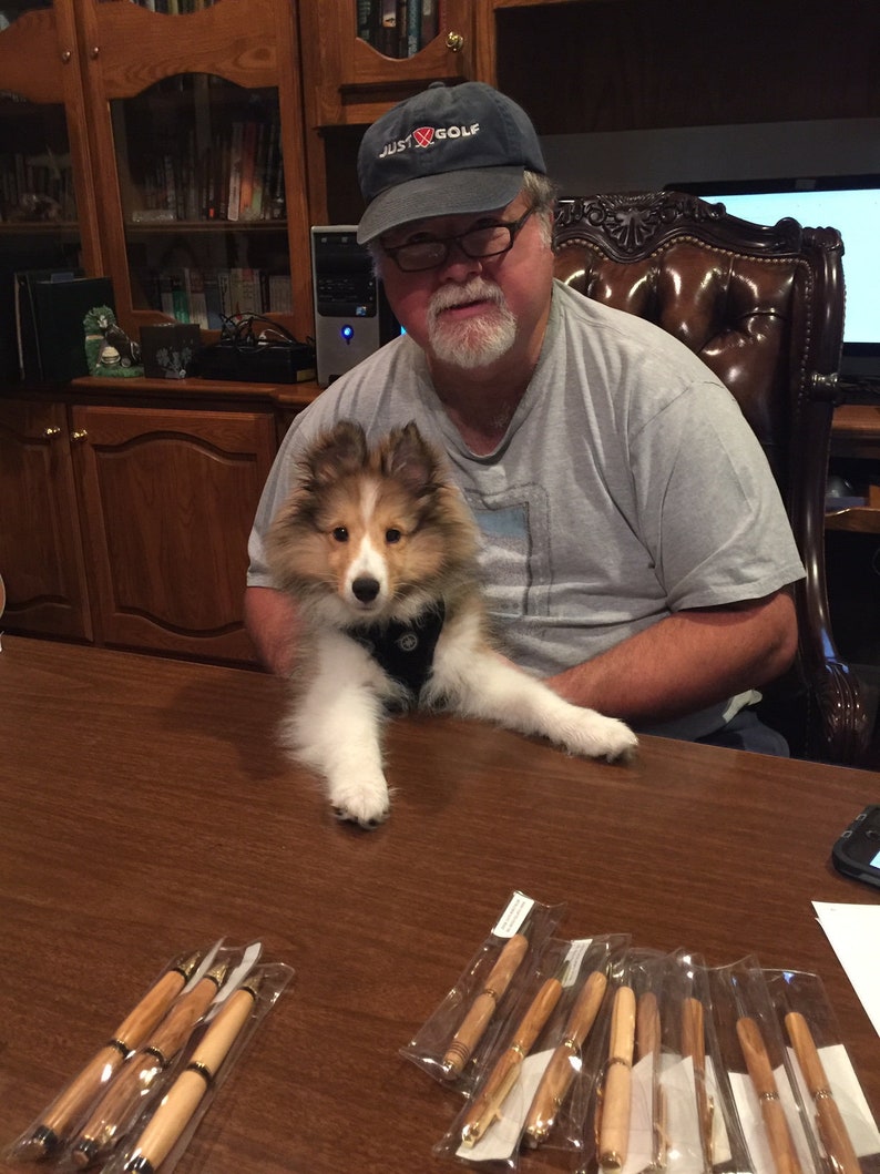 Puede incluir: Una colecci&oacute;n de bol&iacute;grafos de madera con detalles dorados, empaquetados en fundas de pl&aacute;stico transparente, se exhibe sobre una mesa de madera marr&oacute;n oscuro. Un perro peque&ntilde;o, esponjoso, marr&oacute;n y blanco est&aacute; sentado en la mesa. Un hombre con una camiseta gris y una gorra Just Golf est&aacute; en el fondo.