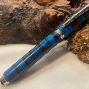 Puede incluir: Un bol&iacute;grafo a rayas azules y negras con clip y detalles plateados. El bol&iacute;grafo tiene una punta redondeada y una banda decorativa cerca de la parte superior. El bol&iacute;grafo est&aacute; hecho de un material liso y est&aacute; sobre un fondo de madera natural.