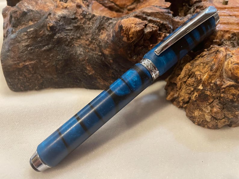 Puede incluir: Un bol&iacute;grafo a rayas azules y negras con clip y detalles plateados. El bol&iacute;grafo tiene una punta redondeada y una banda decorativa cerca de la parte superior. El bol&iacute;grafo est&aacute; hecho de un material liso y est&aacute; sobre un fondo de madera natural.