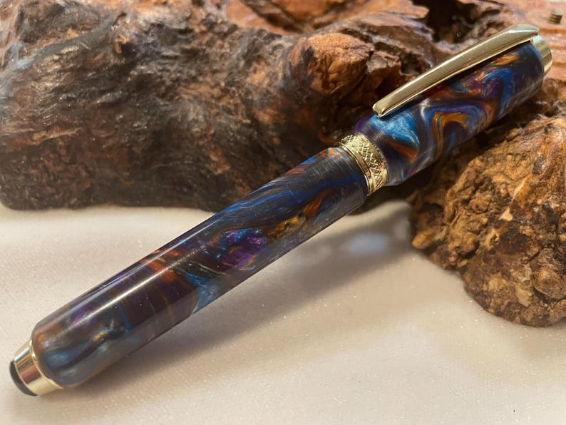 Puede incluir: Una pluma estilogr&aacute;fica artesanal con un patr&oacute;n arremolinado de tonos azules, naranjas y morados. La pluma presenta detalles dorados en el clip y el adorno. La pluma se exhibe sobre una superficie de madera texturizada.
