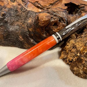 May include: A silver and black pen with a pink and orange ombre resin barrel. The pen has a textured metal cap and a silver tip.