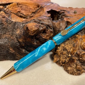 May include: A turquoise and white marbled pen with gold accents. The pen features a gold-colored tennis racket design on the top and a gold-colored tip. The pen is set against a textured, brown wooden background.