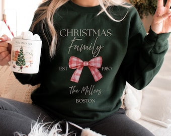 May include: A dark green sweatshirt with the text "CHRISTMAS Family" and a red plaid bow graphic. Below the bow are the words "The Millers" and "BOSTON". A mug with a Christmas scene is held in the left hand.