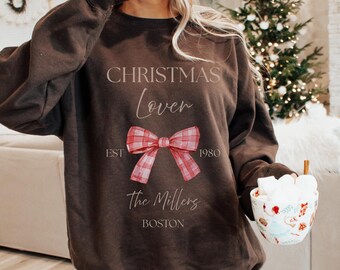 May include: A brown sweatshirt with the text "CHRISTMAS Lover" above a red plaid bow. Below the bow are the words "EST 1980" and "The Millers BOSTON". The sweatshirt is a festive holiday apparel item.