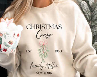 May include: Cream-colored sweatshirt with the text "CHRISTMAS Crew" in black script. Below the text is a sprig of mistletoe with a pink bow. The sweatshirt also includes the text "EST 1980" and "Family Miller NEW YORK".