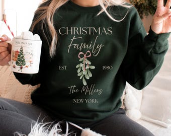 May include: Dark green sweatshirt with the text "CHRISTMAS Family" and "EST 1980" with a mistletoe graphic. Below the text is "The Millers NEW YORK". A white mug with a Christmas scene is held in the left hand.