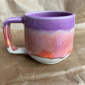 May include: A handmade ceramic mug with a unique color gradient. The top is a deep purple, transitioning to a coral and orange midsection, and a speckled white base. The handle mirrors the color scheme. A functional and artistic piece.