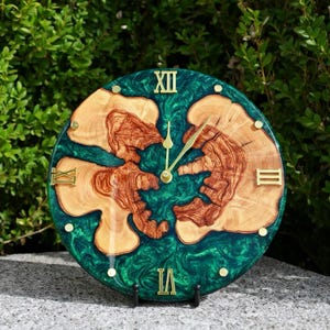 May include: Round wall clock with a green epoxy resin background and natural wood grain accents. The clock face features gold Roman numerals and hands. The clock is displayed on a black stand.