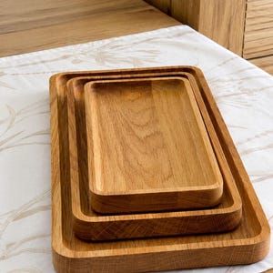 Könnte beinhalten: Ein Set aus drei ineinander gestapelten rechteckigen Holztabletts. Jedes Tablett hat eine hellbraune Oberfläche mit natürlicher Holzmaserung, erhöhten Rändern und abgerundeten Ecken, was für ein glattes und einheitliches Erscheinungsbild sorgt.