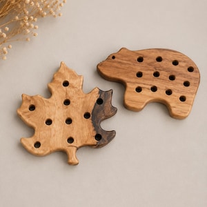 May include: Two wooden objects shaped like a maple leaf and a bear, both featuring a smooth finish and multiple circular holes drilled through the surface. The maple leaf displays a natural wood grain with a dark brown section, while the bear is light brown.