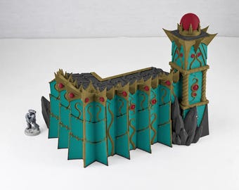 Puede incluir: Una estructura de fortificación en miniatura de color turquesa y dorado con una torre alta con una esfera roja en la parte superior, adornos decorativos y detalles puntiagudos. Una pequeña figura gris sin pintar está al lado.