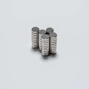 May include: A collection of small, circular, silver-coloured metal magnets arranged in four vertical stacks. Each stack consists of multiple thin, flat, disc-shaped magnets with a smooth, reflective surface, creating a uniform and metallic appearance.