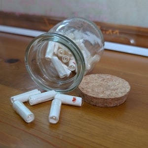 Op de afbeelding: Een heldere glazen pot met een kurken deksel, die op zijn kant ligt en kleine opgerolde papiertjes verspreidt. Sommige papiertjes liggen verspreid rond de pot. De pot staat op een houten oppervlak.