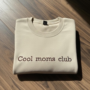 Pu&ograve; includere: Una felpa beige con la scritta ricamata "Cool moms club" in marrone. La felpa &egrave; piegata su una superficie di legno. La felpa girocollo &egrave; di un colore neutro.