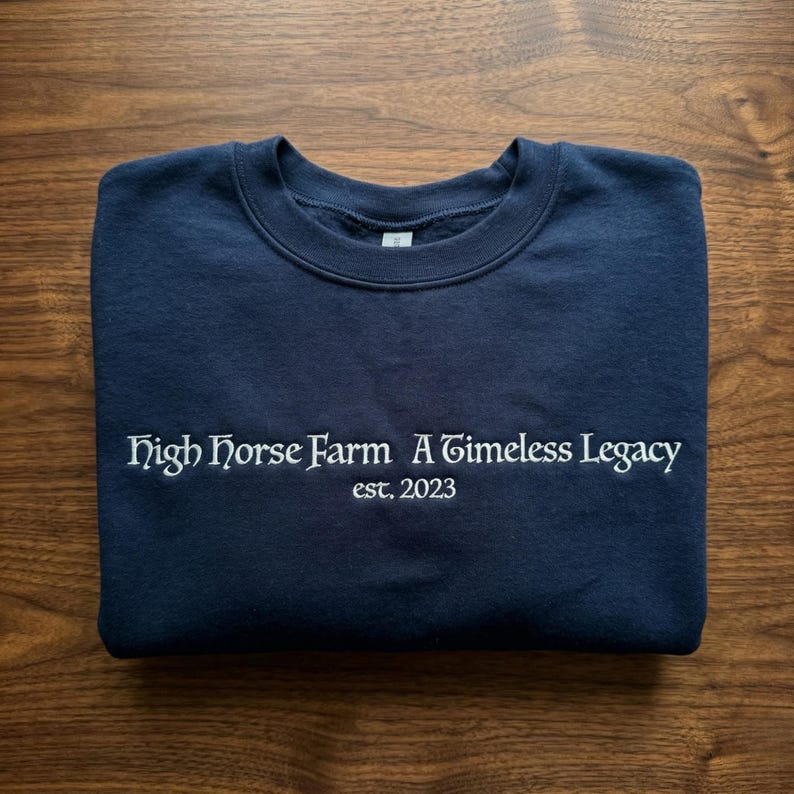 Pu&ograve; includere: Una felpa blu navy con la scritta bianca "High Horse Farm A Timeless Legacy est. 2023". La felpa &egrave; piegata su una superficie di legno.