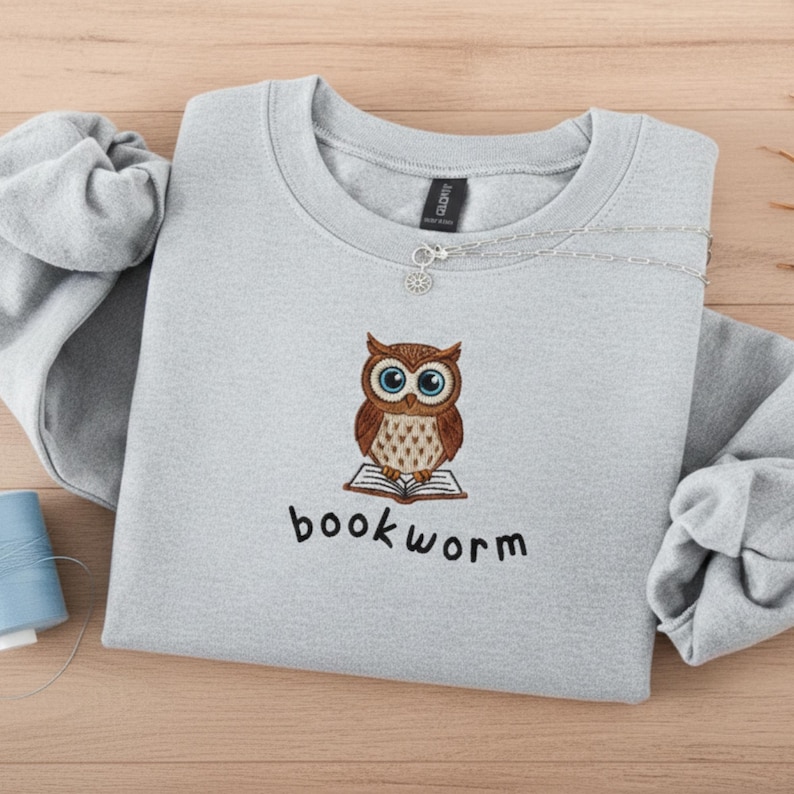 Pu&ograve; includere: Una felpa grigio chiaro con un ricamo a forma di gufo. Il gufo &egrave; marrone con occhi blu e siede su un libro aperto. La parola "bookworm" &egrave; ricamata sotto. Una collana d'argento &egrave; drappeggiata sulla felpa.