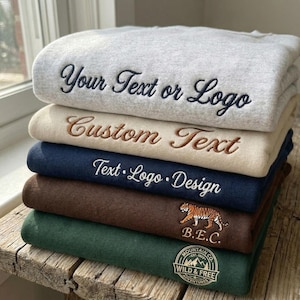 Pu&ograve; includere: Una pila di felpe ricamate in vari colori: grigio, beige, blu navy, marrone e verde. Ogni felpa presenta testo o loghi personalizzati in colori di filo a contrasto. Le due felpe inferiori hanno una grafica di tigre e un logo di montagna.