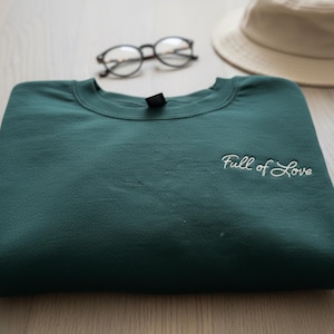 Pu&ograve; includere: Una felpa verde scuro piegata con la scritta ricamata "Full of Love" in corsivo bianco. Un paio di occhiali rotondi e un cappello da pescatore beige sono visibili sullo sfondo.