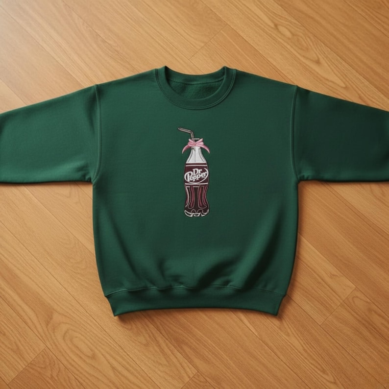 Pu&ograve; includere: Una felpa verde scuro con un design ricamato di una bottiglia di Dr. Pepper. La bottiglia &egrave; raffigurata con un fiocco rosa, una cannuccia e il logo Dr. Pepper. La felpa ha un girocollo e maniche lunghe.