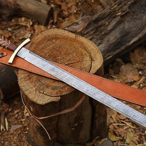 May include: A long, ornate sword with a patterned blade and a leather sheath. The hilt features a gold-colored crossguard and a brown and tan handle. The sword rests on a wooden stump, with a backdrop of leaves and logs.