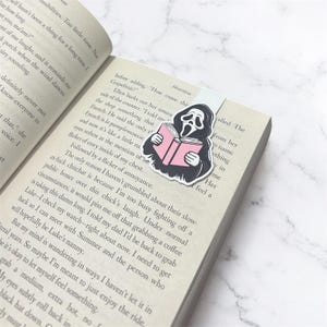 Può includere: Un segnalibro magnetico con un'illustrazione in bianco e nero di una figura incappucciata con una maschera bianca, simile al personaggio Ghostface, che tiene in mano un libro rosa aperto. Il segnalibro è inserito tra le pagine di un libro.