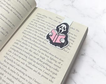 May include: A rectangular paper bookmark featuring a graphic illustration of a black-hooded figure with a white mask reading an open pink book. The bookmark is placed inside an open book with printed text on the pages.