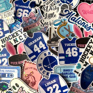 May include: A collection of holographic stickers featuring hockey-themed designs, including blue jerseys with names like Graham 44, Tucker 46, and Logan 22, hockey pucks, sticks, skates, cassette tapes, books, and text such as Dirty Little Secrets.