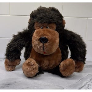 May include: A plush gorilla toy featuring dark brown shaggy fur on the head and arms, with a lighter tan face, chest, and feet. The stuffed animal has a black plastic nose, small dark eyes, and a tiny red accent near the mouth area.
