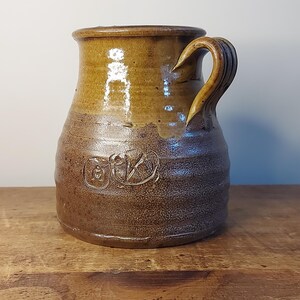 May include: A brown ceramic jug with a single handle and a brown glaze. The jug has a unique design with a raised circle and a cross inside the circle.