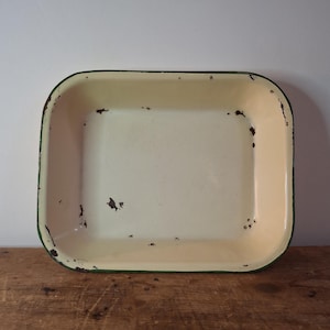 May include: Cream-coloured rectangular enamelware tray with a green trim. The tray has a distressed appearance with visible wear and tear, including chipped paint and rust spots, adding to its vintage charm. The tray is on a wooden surface.