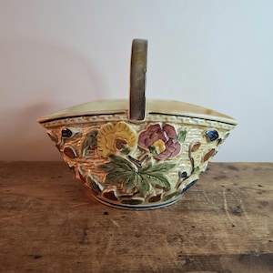May include: A ceramic basket with a handle, featuring a textured, woven design. The basket is adorned with embossed floral and leaf patterns in yellow, purple, and green. The handle is a dark brown colour. The basket sits on a wooden surface.