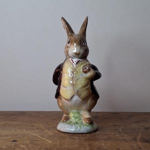 May include: A ceramic rabbit figurine, wearing a yellow waistcoat and brown jacket, holding a pocket watch. The rabbit has brown fur and long ears. The figurine stands on a small patch of green grass.