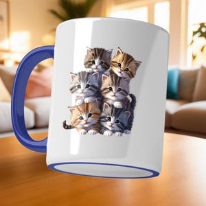 May include: White ceramic mug with a blue handle and rim. The mug features a stack of six adorable kittens in various colors, including brown, gray, and orange. The kittens have detailed fur and are smiling. The mug is sitting on a wooden surface.
