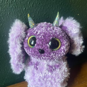 May include: A plush, lavender creature with large, round eyes and glittery horns. The handmade toy has long, fluffy ears and a soft, textured body. The creature's eyes are black with gold accents, and it has a small black nose.