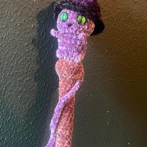 May include: A handmade purple plush witch doll with green eyes and a black hat. The doll is attached to a brown, textured staff. The staff has a purple, twisted accent. The doll is a whimsical, Halloween-themed item.