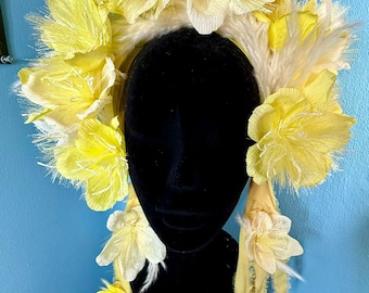 May include: A decorative headpiece featuring large yellow paper flowers and white feathers. The headpiece is displayed on a black mannequin head. The base of the headpiece has a yellow ribbon with flowers. The text "Mango Rose Emporium" is visible on the base.