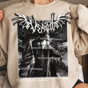 Peut inclure: Un sweat-shirt beige avec un motif graphique en noir et blanc. Le motif comprend le mot "Desecrate" dans une police stylisée, ainsi qu'une illustration détaillée d'une figure avec une épée. Le texte "ALYARTSDESIGN" est également visible.
