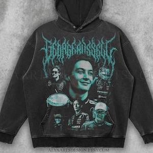 May include: Black hooded sweatshirt featuring a portrait of George Russell with the text "GEORGE RUSSELL" in teal. The design includes images of Russell and other Formula 1 elements. The sweatshirt has a front pocket and a drawstring hood.