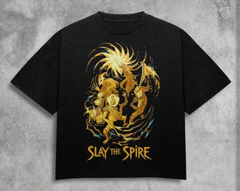May include: Black t-shirt featuring a golden illustration of figures and the text "SLAY THE SPIRE". The design includes stylized characters and a sunburst element. The shirt is made of a soft material.