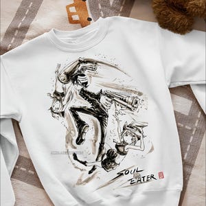 May include: White sweatshirt featuring a black and white anime-style illustration of characters with a gun. The text "SOUL EATER" is printed in black ink. The sweatshirt is on a patterned surface.