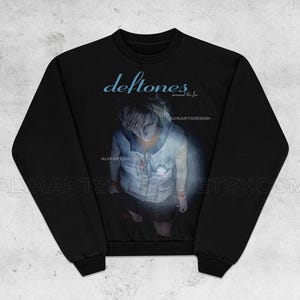 May include: Black sweatshirt featuring the band name "Deftones" in light blue, with the text "around the fur" below. The graphic shows a person in a white vest and dark skirt. The Etsy shop name "ALYAARTSDESIGN" is also visible.